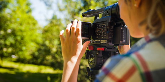 Photo: Close-up of a female Student who is holding a VJ camera to her face. We can see her left ear, shoulder and part of the face, her left hand is adjusting the camera. Background: trees and grass.