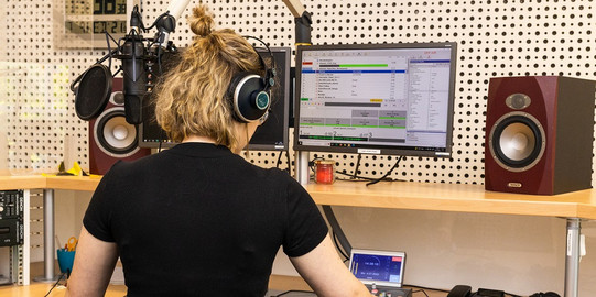 Photo: A female student is standig in the eldoradio* radio station, her back turned towards the camera. She is occupied with adjusting studio technology.