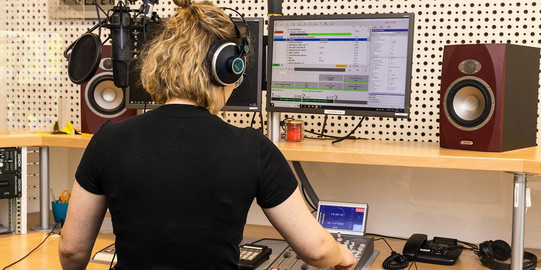 Photo: A female student is standig in the eldoradio* radio station, her back turned towards the camera. She is occupied with adjusting studio technology.