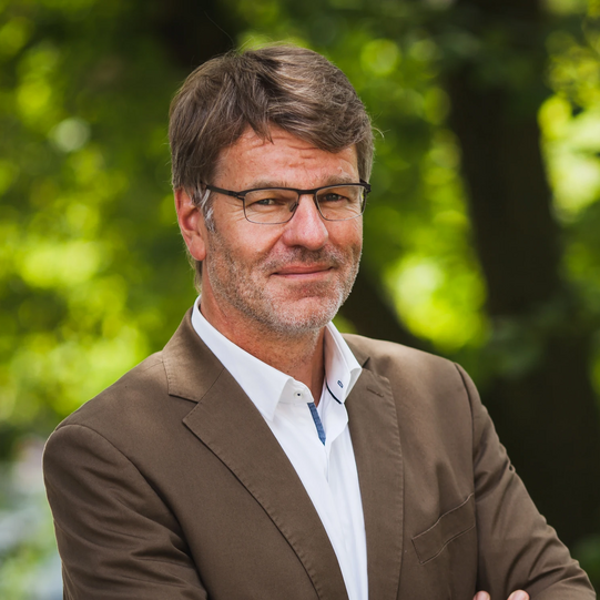 Photo: Profile picture of Prof. Dr. Henrik Müller, his arms are crossed in front of the chest, he is wearing glasses, a whithe shirt and brown jacket. Background: trees.
