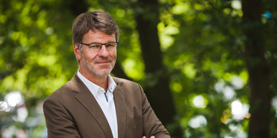 Photo: Profile picture of Prof. Dr. Henrik Müller, his arms are crossed in front of the chest, he is wearing glasses, a whithe shirt and brown jacket. Background: trees.