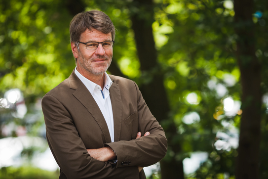 Photo: Profile picture of Prof. Dr. Henrik Müller, his arms are crossed in front of the chest, he is wearing glasses, a whithe shirt and brown jacket. Background: trees.