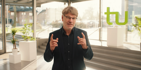 Photo: Prof. Dr. Henrik Müller, wearing a black shirt and glasses, is standing in a very light room, green TU letters are in the background. Müller raises both hands and index fingers in front of him and looks straight into the camera.