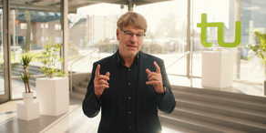 Photo: Prof. Dr. Henrik Müller, wearing a black shirt and glasses, is standing in a very light room, green TU letters are in the background. Müller raises both hands and index fingers in front of him and looks straight into the camera.