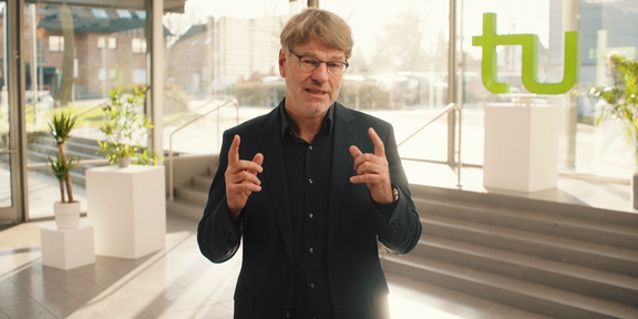 Photo: Prof. Dr. Henrik Müller, wearing a black shirt and glasses, is standing in a very light room, green TU letters are in the background. Müller raises both hands and index fingers in front of him and looks straight into the camera.