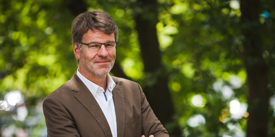 Photo: Profile picture of Prof. Dr. Henrik Müller, his arms are crossed in front of the chest, he is wearing glasses, a whithe shirt and brown jacket. Background: trees.