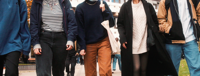 5 students, male and female, are walking on the campus in front of EF 50 building, they are happily chatting, the brownish leaves on the trees and the student's clothing suggest that it's autumn