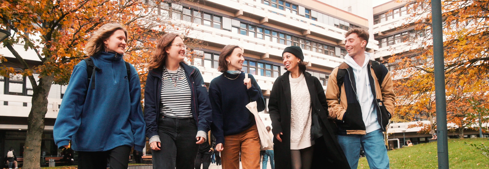 5 students, male and female, are walking on the campus in front of EF 50 building, they are happily chatting, the brownish leaves on the trees and the student's clothing suggest that it's autumn