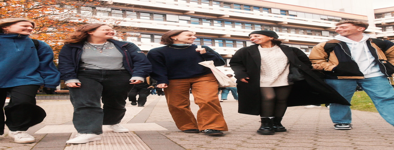 5 students, male and female, are walking on the campus in front of EF 50 building, they are happily chatting, the brownish leaves on the trees and the student's clothing suggest that it's autumn