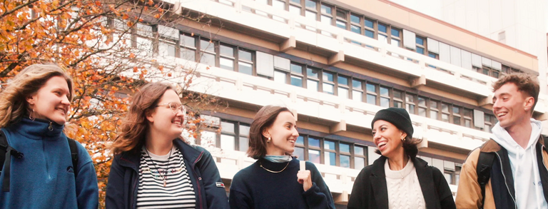Header image IJ students autumn EF50 5 students, male and female, are walking on the campus in front of EF 50 building, they are happily chatting, the brownish leaves on the trees and the student's clothing suggest that it's autumn