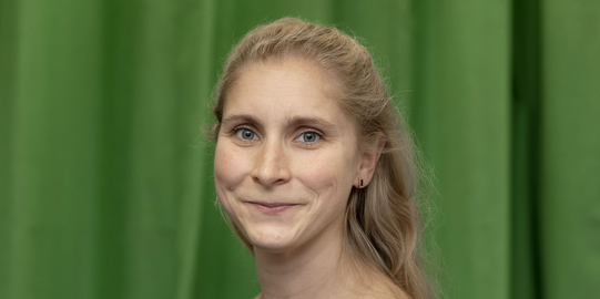 Photo: Profile picture of Lena Marie Hufnagel. She is wearing a dark blue dress with with large floral print and has shoulder-long blond hair. Background: green-screen.