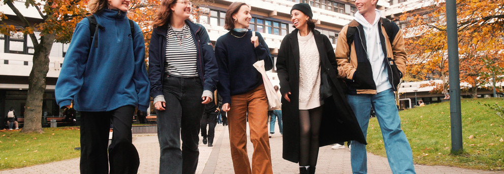 Photo: 5 students, male and female, are walking beside each other on the campus, their faces are turned toward each other. Background: trees with brown-yellow leaves and EF50 building.