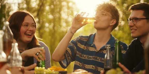 Photo: 3 students are having a cold drink in the late afternoon sun. Background: sun is shining through tree leaves.