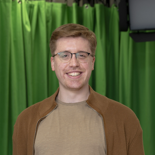 Photo: Profile picture of Nico Hornig. He wears glasses, a khaki T-Shirt and a brown cardigan. Background: green screen.