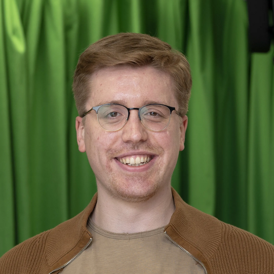 Photo: Profile picture of Nico Hornig. He wears glasses, a khaki T-Shirt and a brown cardigan. Background: green screen.