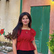 Photo: Profile picture of student Mariam Fayz, she is wearing long black pants and a red shirt. She is standing on a paved road with a palm tree in the background