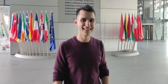Photo: Research associate Juan Pedro Díaz Martínez is standing in the European Parliament, flags from EU member countries are to his right and left side, EU name plate in all EU languages in the background.