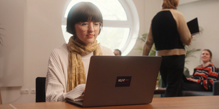 Photo: Girl with short dark hair, glasses and white sweatshirt is sitting at a desk, typing on a laptop