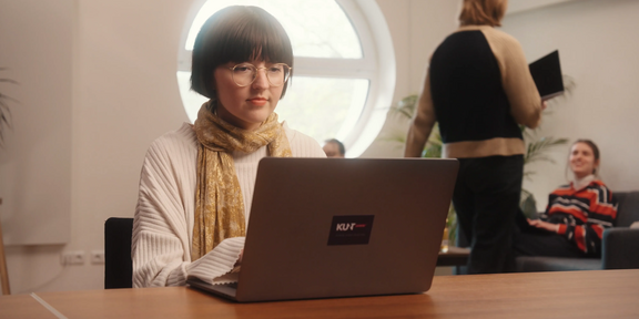 Photo: Girl with short dark hair, glasses and white sweatshirt is sitting at a desk, typing on a laptop