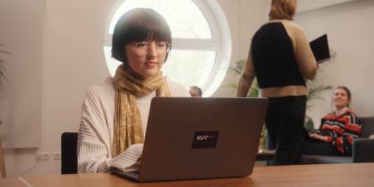 Photo: Girl with short dark hair, glasses and white sweatshirt is sitting at a desk, typing on a laptop