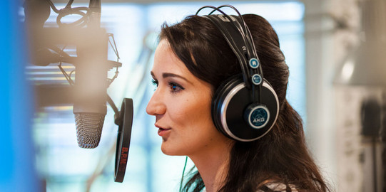 Photo: Female student with dark, shoulder long hair and headphones, is speaking into a microphone at a radio station.