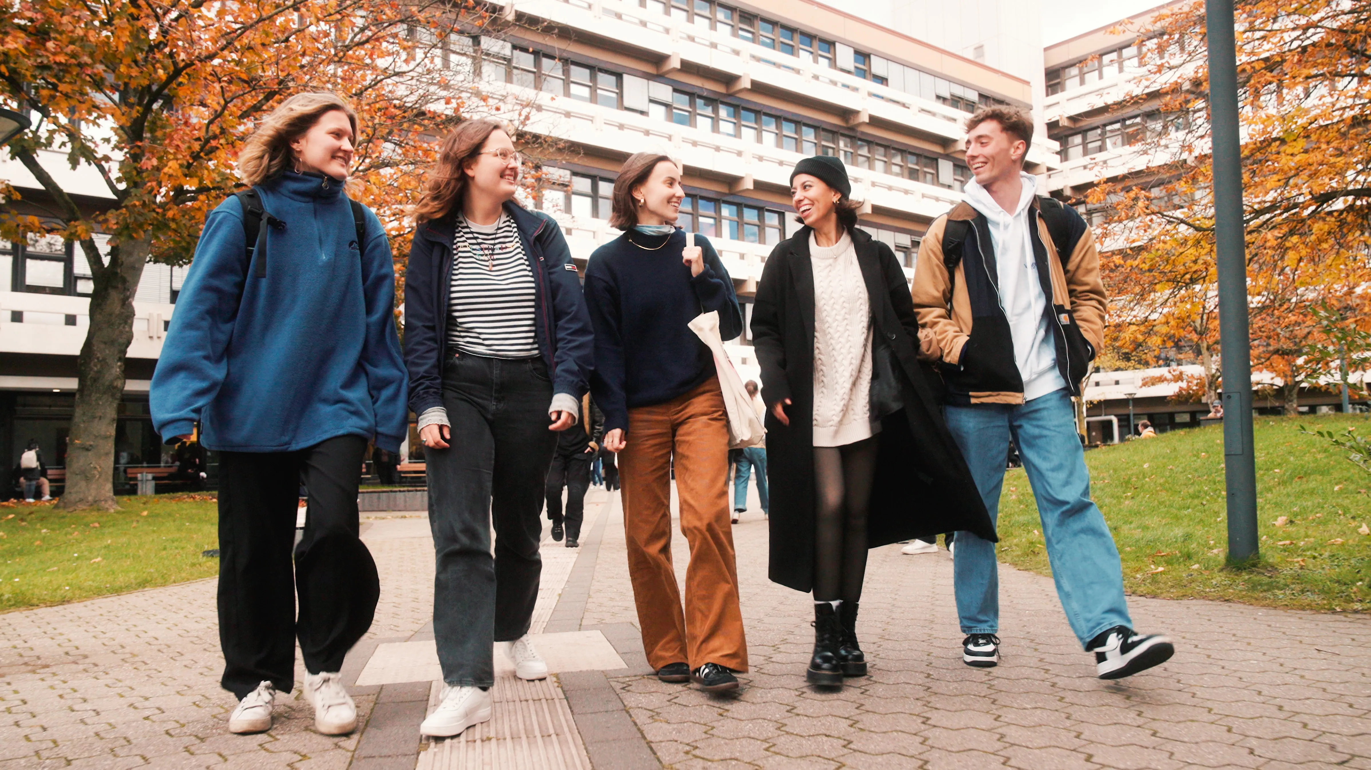 5 students, male and female, are walking on the campus in front of EF 50 building, they are happily chatting, the brownish leaves on the trees and the student's clothing suggest that it's autumn