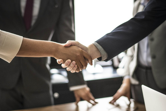 Two people shake hands. The handshake is shown in detail, in the background are two people in suits.